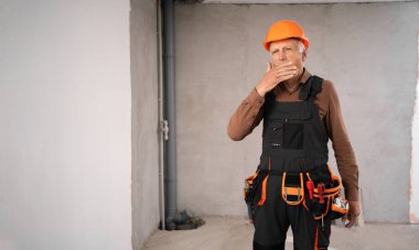 Yorgun, uykulu emekliler inşaat şantiyesi çalışanı koruyucu takım elbiseli ve iş yerinde iş tulumu giyiyor. Boşluğu kopyala