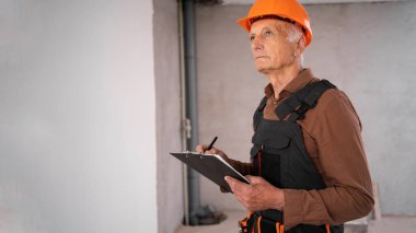 Miğferli yaşlı inşaat işçisi yeni inşaat alanındaki inşaat çalışmalarını kontrol ediyor. Tecrübe kavramı, kalite kontrolü, denetim ve ev geliştirme.