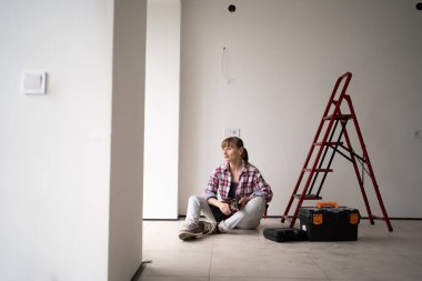 İşçi ya da tamirci. Yeni ev ya da dairede alet kemeri takan bir kadın merdiven ve alet çantasının yanında oturuyor. Yenileme ve onarım kavramı