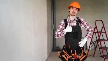 İnşaat alanında çalışan, baş parmağını kaldıran genç bir inşaatçının portresi. Pankart. Boşluğu kopyala