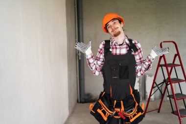 Miğferli ve alet kemerli miğferli bir adam inşaat sahasında duruyor. Kafası karışmış, kolları özür diliyor. Ev yenileme ve müteahhit sorumluluğu sırasındaki belirsizlik kavramı.