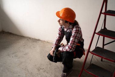 İnşaatçı kasketli bir adam yenilenmiş odada oturuyor ve elektrikli matkabı çalışmaya hazır tutuyor. İnşaat alanı, inşaat süreci, ev yenileme ve çalışan insanlar kavramı.