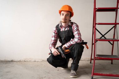 Erkek inşaat işçisinin portresi merdivenin yanında oturuyor ve ev yenileme alanında matkap tutuyor. İnşaat alanı, inşaat süreci, ev onarımı ve çalışan insanlar kavramı.