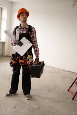Erkek işçi dairede. Kasketli ve işçi üniformalı bir işçi boş bir odada elinde alet çantası, dosya ve planlarla dikiliyor. Tamirat, inşaat ve yenileme konsepti