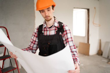 Turuncu miğferli bir erkek müteahhit planlar okuyor, ev yenileme projesini gözden geçiriyor, inşaat sürecinde evde duruyor, ev onarımı, planlama, mühendislik, geliştirme...