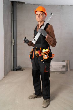 Duvarı tutan kemerli yaşlı inşaatçı kameraya kibarca bakıyor. Ev yapımı ve yenileme konsepti