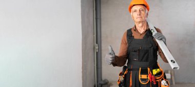 İnşaat işçisi, ruhsal seviyesi ve aletleri olan yaşlı bir adam. İnşaat sahasında duruyor. El hareketi sınıfının yükseldiğini gösteriyor. Boşluğu kopyala