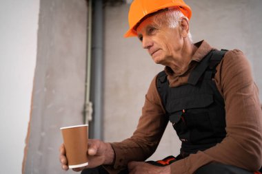 Üniformalı yaşlı inşaatçı şantiyede oturmuş kahve içiyor. inşaat ve endüstri konsepti.