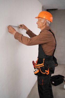 Yumuşak metre kullanarak tamamlanmamış bir evdeki duvar ölçümleri sırasında profesyonel bir yaşlı inşaatçı inşa etmek. Yan görünüm