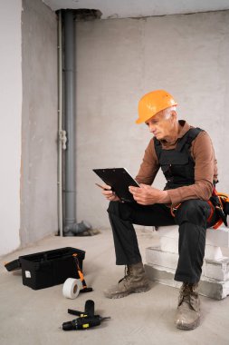 İnşaat mühendisi, pano okuma listesi olan yaşlı bir adam inşaat alanında teftiş notları hazırlıyor. Onarım, yenileme ve inşaat konsepti