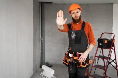 Sakallı bir tamirci ya da miğferli bir müteahhit ve tulumlu ciddi ciddi üzgün bir kamera. Durma hareketini elinde avucunda inşaat ekipmanlarıyla gösteriyor. Yapı ve onarım kavramı