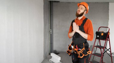 Koruyucu miğferli ve tulumlu sakallı müteahhit el ele dua ediyor. İnşaat malzemeleriyle odada iyi şanslar diliyor. İnşaat ve onarım konsepti. Pankart