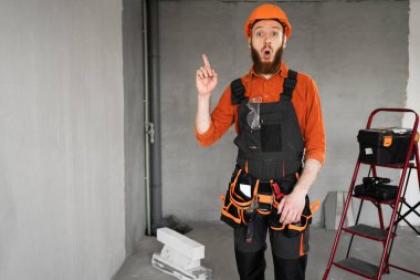 Koruyucu miğferli ve tulumlu sakallı müteahhit odada inşaat ekipmanlarıyla harika bir fikir olduğunu gösteriyor. Yapı ve onarım kavramı