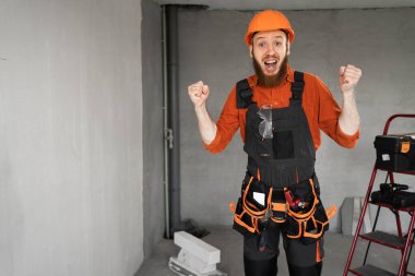 Sakallı bir tamirci ya da miğferli bir müteahhit ve çok mutlu ve heyecanlı, kazanan jestler yapıyor, kolları havada odada. Kutlama, inşaat ve onarım konsepti