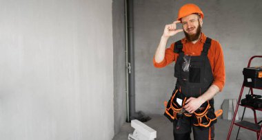 Tamirci ya da miğferli ve tulumlu müteahhit ellerini bina ekipmanlarıyla odada gezdirirken küçük boy jest gösteriyor. Yapı ve onarım kavramı