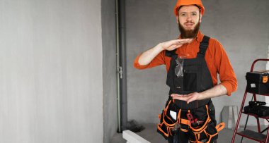 Tamirci ya da miğferli ve tulumlu müteahhit elleri inşaat malzemeleriyle dolu odada büyük bir jest gösteriyor. Yapı ve onarım kavramı