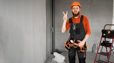 İnşaatçı inşaat işçisi kasketli, gülümseyen bir şekilde dairede barış işareti yapıyor. Boşluğu kopyala