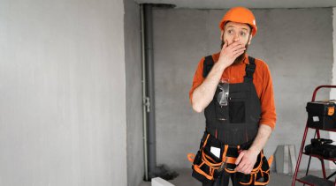 Şok olmuş müteahhit turuncu miğfer giyer, şaşkınlığını ifade eder, ağzını kapatır, güvensizlik ve sürpriz duygular gösterir, onarım işlemleri odasında durur. Pankart. Boşluğu kopyala