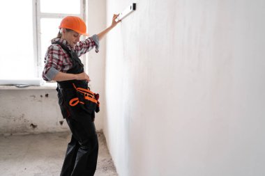 Koruyucu kask takan kadın inşaat işçisi veya inşaatçı, yenileme veya inşaat çalışmaları sırasında ölçüm duvarı olarak ruh seviyesini kullanır. Pankart. Boşluğu kopyala