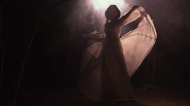 Dolunay altında uzun beyaz elbiseler içinde dans eden genç bir kadının silueti gizemli bir ormanda zarif bir hareket doğal güzellik, büyülü bir atmosfer ve duygusal ifade sergiliyor..