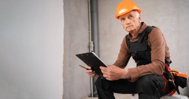 Üst düzey inşaat işçisi elinde pano, oturup dikkatlice bakıyor. Ev tadilatı, apartman tadilatı ve inşaat süreci.