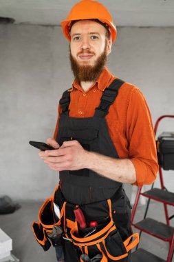 Akıllı telefonlu ve kemerli aletleri olan genç sakallı erkek müteahhit yeni evde ayakta duruyor. Teknoloji, onarım, inşaat ve inşa kavramı.