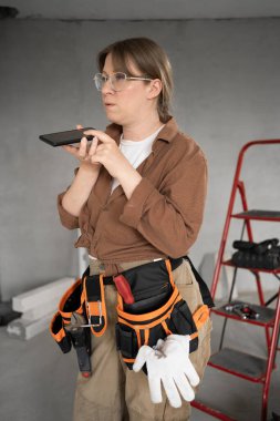 Modern teknolojileri kullanan kadın ya da tamirci. Alet kemerindeki kadın işçi akıllı telefondan sesli mesaj kaydediyor, inşaat alanında sanal asistanlık yapıyor. Boşluğu kopyala