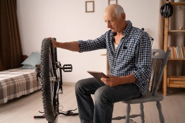 Rahat bir dairede oturan yaşlı beyaz bir adam bisikleti tamir ediyor, tablette video dersleri alıyor. Cihazlar yakında, odaklanmış durumda. Emeklilikte hobiler ve aktif yaşam ilham ve beceri doludur..