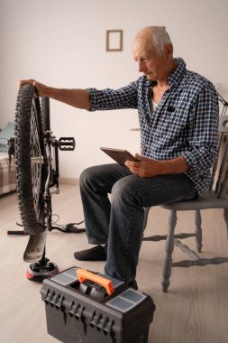 Rahat bir dairede oturan yaşlı beyaz bir adam bisikleti tamir ediyor, tablette video dersleri alıyor. Cihazlar yakında, odaklanmış durumda. Emeklilikte hobiler ve aktif yaşam ilham ve beceri doludur..
