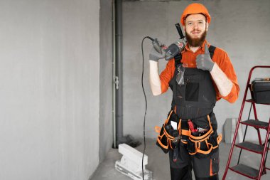 Turuncu kasklı ve tulumlu sakallı tamircinin portresi, inşaat alanında matkap delgisi tutan başparmağı gösteriyor. Boşluğu kopyala