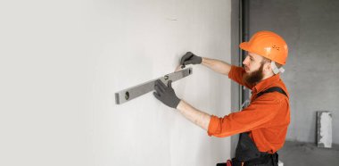 Tamir ve inşaat konsepti. Koruyucu miğferdeki erkek inşaatçı ya da mühendis ruh seviyesiyle duvarı ölçer. Beton duvarın yanındaki evde mimar. Boşluğu kopyala