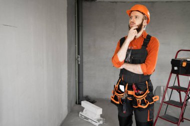 Sakallı bir tamirci ya da miğferli ve tulumlu bir tamirci. Hayal gibi görünüyor. İnşaat malzemeleriyle odada düşünüyor. Yapı ve onarım kavramı