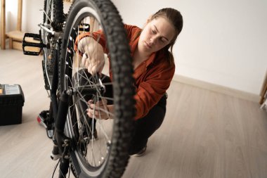 Evde yerde diz çöküp bisikleti tamir eden genç bir kadın, tekerlek somununu sıkmak için ingiliz anahtarı kullanıyor. DIY onarımı, ev bakımı, mekanik ayar ve bisiklet donanımı bakımı kavramı.