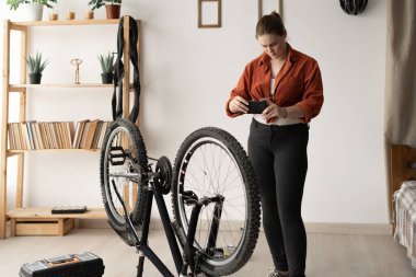 Young woman standing next to her bicycle at home taking a photo of it with her smartphone. DIY concept. Capturing details for reference or sharing.