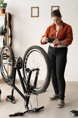A female cyclist stands next to her bicycle in an apartment, taking a photo of it with her phone. Scene of bicycle repair or customization. Documenting the process.