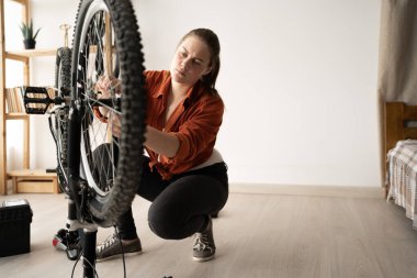 A young Caucasian woman is intently examining the front wheel of her bicycle while doing some home repairs. The atmosphere is DIY and sports equipment maintenance.