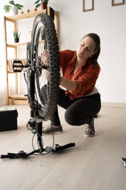 Caucasian girl carefully examines the front wheel of her bicycle in her home workshop. Preparing for a ride or looking for a fault in the parts.