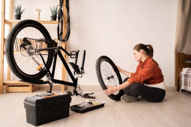 Genç bir bayan bisiklet tamircisi yerde oturur, bisikletinin ön tekerleğini dikkatle inceler. Ev onarımı, hobi.