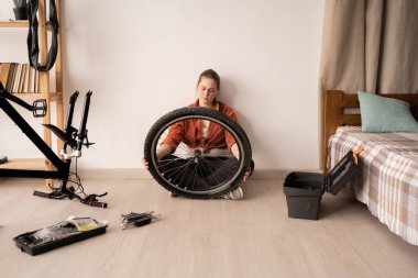 Genç bir bayan yerde oturuyor, dikkatle bisikletin ön tekerleğini inceliyor. Kendin tamir et, hobi. Pankart. Boşluğu kopyala.