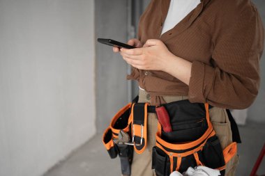female builder with smartphone and working tools on belt, close-up. repair, renovation, construction and building concept
