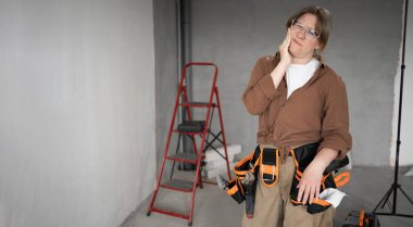 Repairwoman with tool belt in living room interior doing a home renovation, suffering teeth pain. Teeth dental problem concept. Banner. Copy space