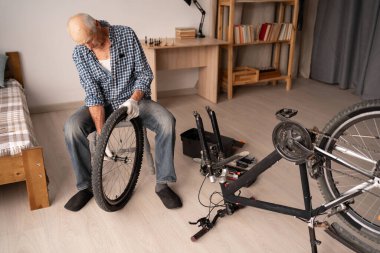 Günlük kıyafetli emekli adam evde, oturma odasının içinde bisiklet tekerleği ayarlıyor. Bisiklet tamiri, bakım onarımı. Bisiklet hobisi kavramı. Yüksek açı