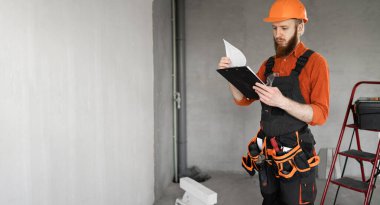 İnşaatçı inşaat işçisi. Alet kemeri ve kaskında inşaat alanında ya da yeni bir evde pano var. Uzayı kopyala Pankart