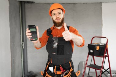 Elektronik ödemeler için POS terminali tutan kasklı gülümseyen bir müteahhit. Boşluğu kopyala