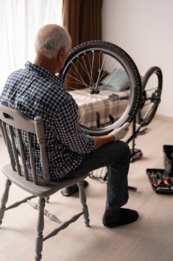 Evde Bisiklet Tamir Ederken Bisiklet Tekerleği Tutan Yaşlı Adam, Sandalyede Otururken. Boşluğu kopyala