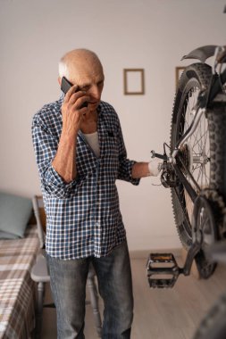 70 yaşında bir adam evde bisiklet tamir ediyor, tamirciye danışmak için telefon kullanıyor. Kıdemli DIY onarımı, mobil destek, ulaşım onarımı ve kapalı mekanik servis kavramı.