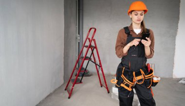 Onarım, inşaat ve iletişim konsepti. Arkaplanda inşaat ekipmanı olan akıllı telefon kullanan kadın işçi ya da müteahhit. Boşluğu kopyala