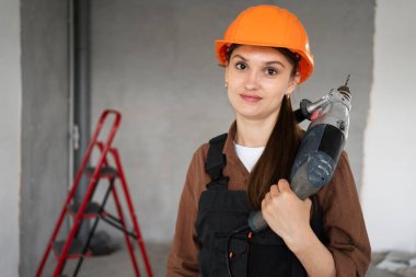 Elektrikli matkap tabancasıyla kameraya bakan mutlu bir kadın inşaatçı. Üniformalı ve kasklı bir zanaatkar. Boşluğu kopyala