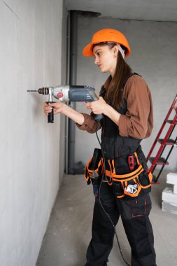 Kasketli ve alet kemerli bir kadın evde matkapla beyaz duvarı deliyor. Boşluğu kopyala