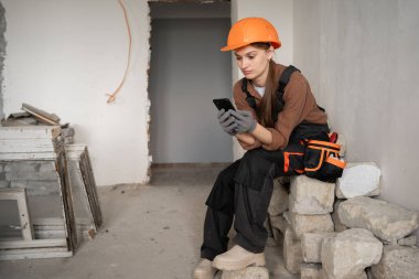 Kadın İnşaatçı, eski binadaki iş molasında akıllı telefon kullanıyor. Boşluğu kopyala